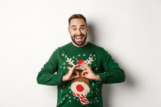 New Year, holidays and celebration. Happy young man making fun of his sweater and smiling, fool around on Christmas party, standing over white background.