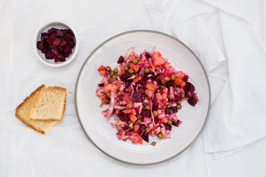 Russian traditional salad with vegetables - vinaigrette on a plate, bread and a bowl of beets on a table on a cloth. Top view