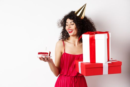 Cheerful smiling woman in red dress and party cone, looking happy at birthday cake, holding gifts, standing on white background. Copy space