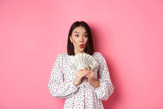 Shopping concept. Excited asian woman holding money, gasping amazed and staring at camera, standing over pink background.