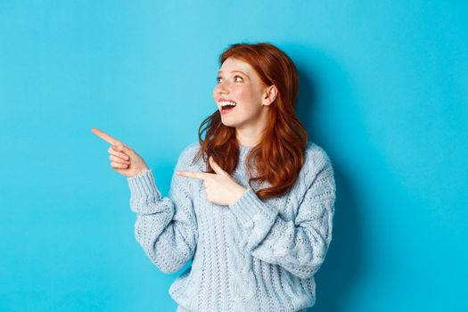 Excited redhead girl in sweater, looking and pointing fingers left, showing promo offer or logo, standing over blue background.