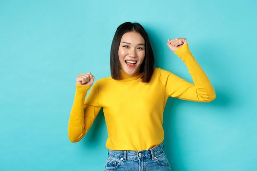Cheerful asian woman partying, having fun and dancing over blue background, triumphing and scream yes with joy, winning prize.