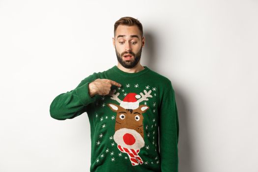 Winter holidays and christmas. Confused guy in sweater pointing at himself, standing puzzled over white background.