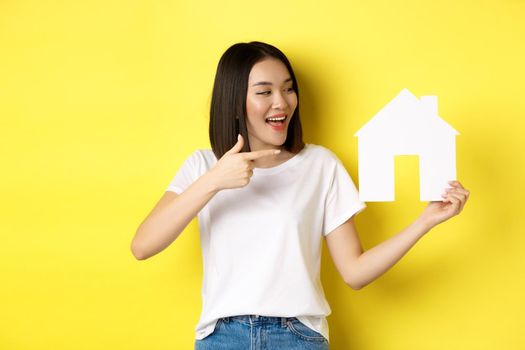 Real estate and insurance concept. Cheerful asian woman smiling, pointing and looking at paper house cutout, recommend agency logo, standing over yellow background.