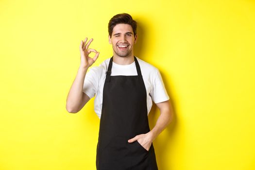 Confident and handsome waiter showing ok sign, wearing black apron and standing against yellow background.