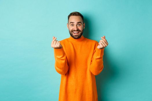 Smiling handsome man showing hearts and looking happy, lucky gesture, standing in orange sweater over light blue background.