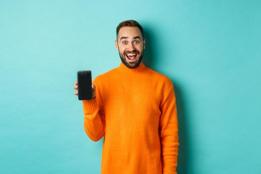 Photo of happy man showing mobile screen, introduce online store, application, standing over turquoise background.