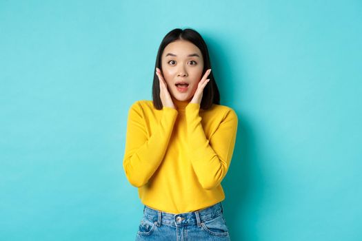 Portrait of surprised asian girl checking out promo, gasping amazed and touching cheeks, staring at camera amazed, blue background.