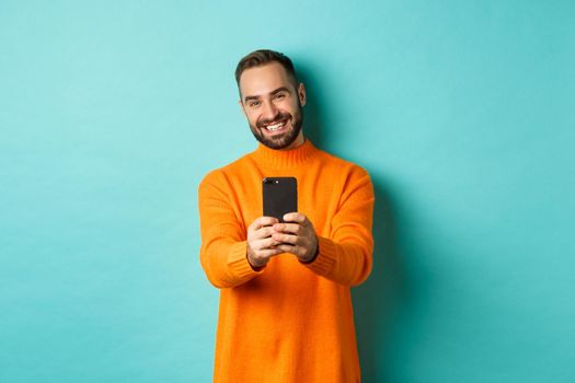 Happy handsome man taking photo on mobile phone, take pictures with smartphone camera, standing over light blue background.