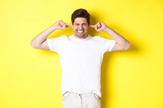 Annoyed man grimacing and shutting ears, complaining on loud noise, standing against yellow background. Copy space