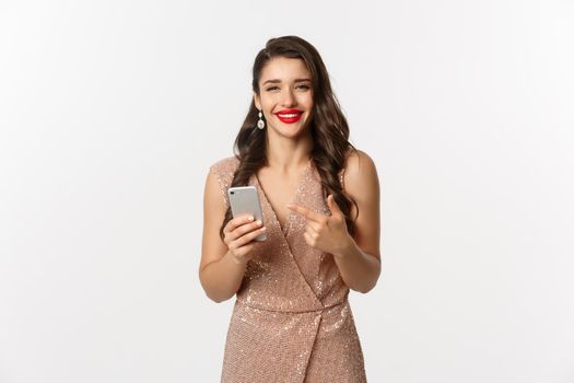 Holidays, online shopping concept. Satisfied good-looking woman in luxury dress and red lipstick, pointing at mobile phone, smiling pleased, white background.