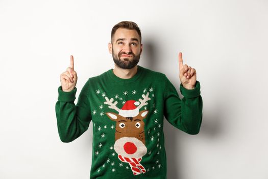 Winter holidays and christmas. Displeased guy looking doubtful at copy space, pointing fingers up and grimacing, standing over white background.
