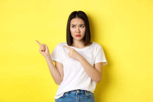 Beauty and fashion concept. Beautiful asian woman in white t-shirt pointing fingers left, standing over yellow background.