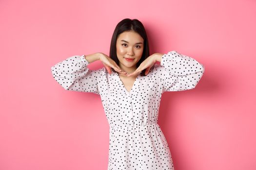 Beautiful asian woman in trendy dress making cute face, holding hands near jawline and gazing coquettish at camera, standing over pink background.