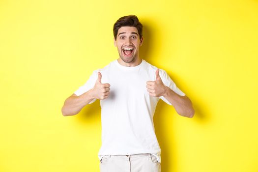 Excited handsome man showing thumbs up, approve and saying yes, standing over yellow background.