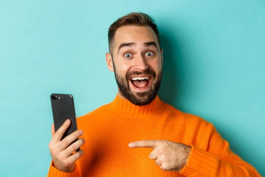 Close-up of impressed man showing something on mobile phone, pointing at smartphone and gasping amazed, standing in orange sweater over light blue background.