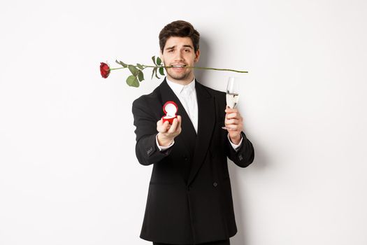 Romantic young man in suit making a proposal, holding rose in teeth and glass of champagne, showing engagement ring, asking to marry him, standing against white background.