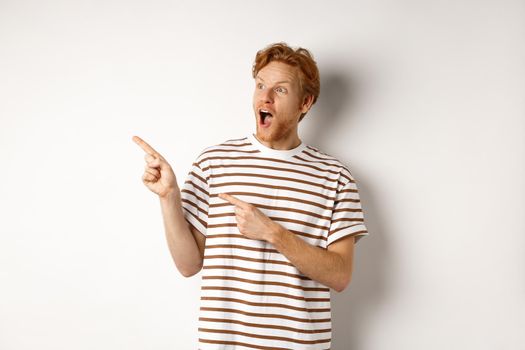 Check it out. Amazed redhead guy pointing and looking left at promotion banner, standing over white background.