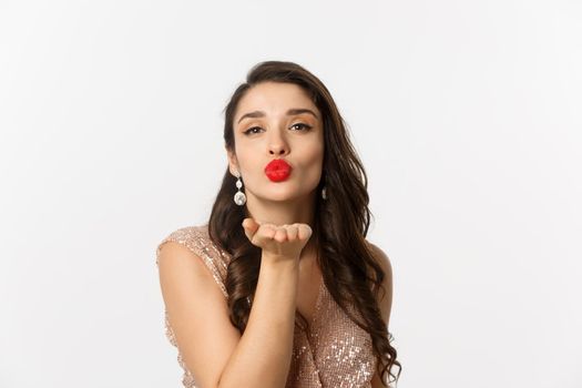 Concept of New Year celebration and winter holidays. Close-up of sensual young woman in dress, pucker lips and blowing air kiss at camera, white background.
