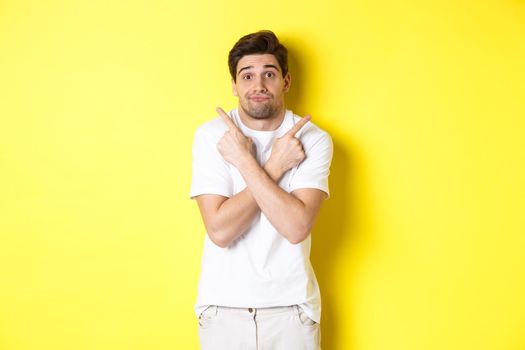 Indecisive man pointing fingers sideways, struggling to make decision, asking advice, standing over yellow background.