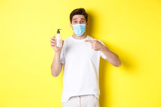 Concept of covid-19, quarantine and lifestyle. Excited guy in medical mask showing good hand sanitizer, pointing finger at antiseptic, standing over yellow background.