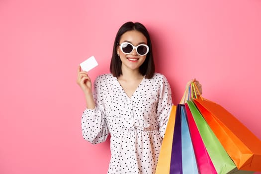 Beautiful asian woman in sunglasses going shopping, holding bags and showing credit card, standing over pink background.