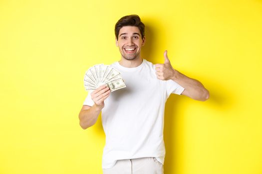 Satisfied buyer man showing thumb-up and holding money, shopping with cash, standing over yellow background.