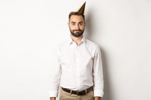 Celebration and holidays. Gloomy guy in birthday hat standing awkward against white background, feeling unamused.