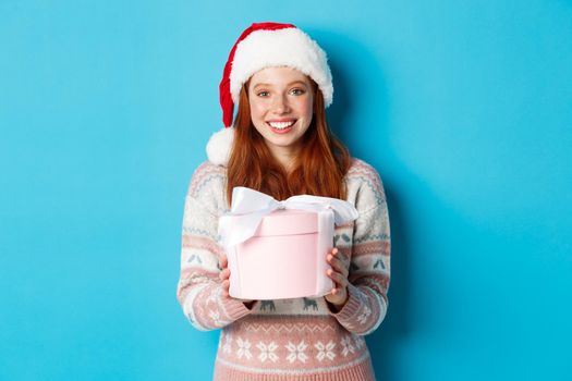 Winter and celebration concept. Adorable redhead girl giving you a present, wishing merry christmas, standing i santa hat against blue background.