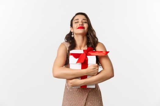 Holidays, celebration concept. Happy and thankful woman hugging Christmas gift, wearing luxury dress, receiving New Year present, standing over white background.