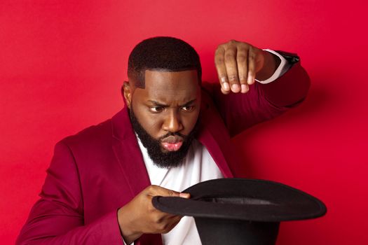 Close-up of charismatic Black male magician perform a trick with his hat, performing on christmas party, standing over red background.