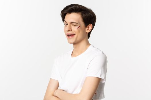 People, lgbtq and beauty concept. Beautiful cheeky gay man winking at camera and smiling, standing with glitter on face against white background.