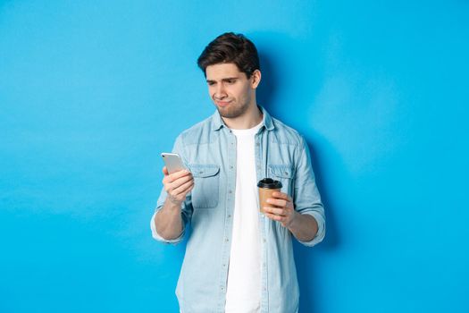 Skeptical and disappointed guy reading upsetting message on phone, holding coffee cup, standing over blue background.