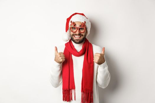 Christmas, New Year and celebration concept. Excited man in Santa hat and party glasses, showing thumbs up in approval, smiling satisfied, white background.