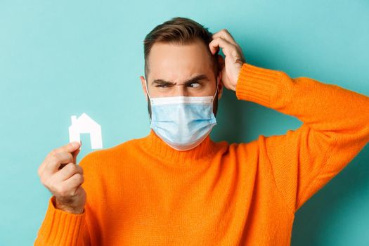 Real estate and coronavirus pandemic concept. Confused man in face mask scartching head while looking at small paper house, wearing orange sweater near light blue studio background.