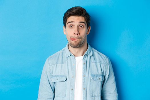 Close-up of young man making funny expressions, showing tongue and looking at camera, standing over blue background.