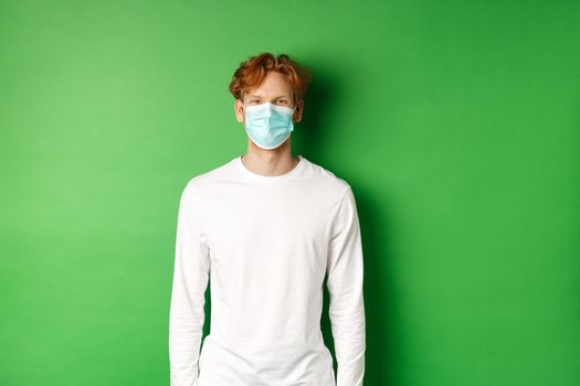 Covid-19, social distancing and lifestyle concept. Young redhead man wearing face mask during coronavirus pandemic, standing over green background.