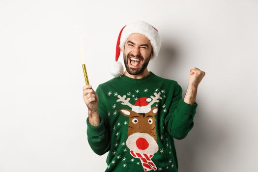 Christmas party and holidays concept. Happy young man celebrating New Year xmas, holding sparkler and looking excited, standing over white background.