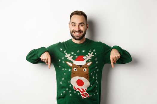 Winter holidays and christmas. Handsome smiling man with beard, pointing fingers down and showing promo offer, wearing green sweater, standing over white background.