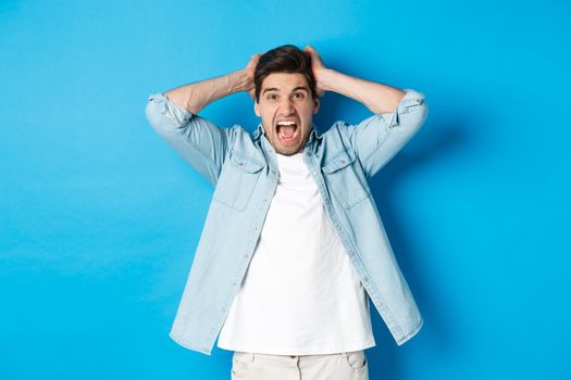 Frustrated guy losing, yelling displeased and looking at camera, holding hands on head, standing over blue background.