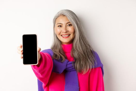 Online shopping. Close up of stylish asian senior woman extending hand with mobile phone, showing blank smartphone screen and smiling, standing over white background.