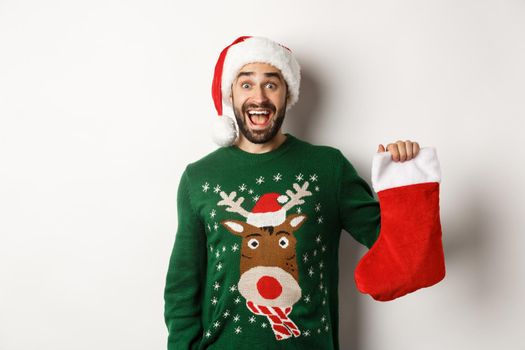 Xmas and winter holidays concept. Happy man got a gift in Christmas sock, looking excited, standing in Santa hat against white background.