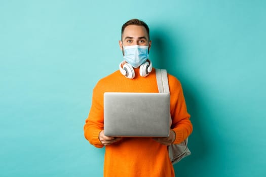 Young man in face mask using laptop and staring happy at camera, standing over light blue background.