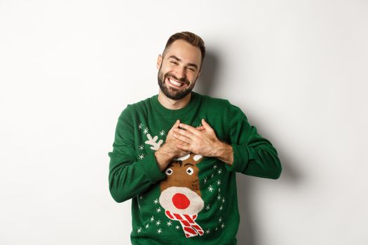 New year celebration and winter holidays concept. Happy young man looking grateful, holding hands on heart and thanking for christmas gift, standing in funny sweater.