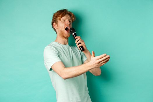 Passionate redhead man in t-shirt singing serenade with microphone, looking aside at karaoke and gesturing, standing over mint background.