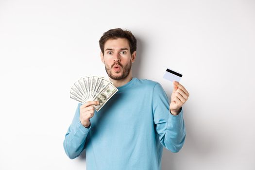 Surprised young man showing money in dollars and plastic credit card, gasping and saying wow with amazed face, standing on white background.