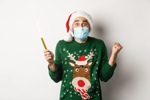 Concept of covid-19 and Christmas holidays. Happy young man in face mask celebrating Christmas, holding party sparkler and rejoicing, standing over white background.