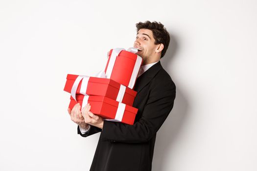 Concept of christmas holidays, celebration and lifestyle. Image of excited handsome man buying gifts for new year, standing happy over white background in black suit.