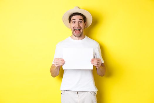 Excited tourist showing your logo or sign on blank piece of paper, smiling amazed, standing against yellow background.