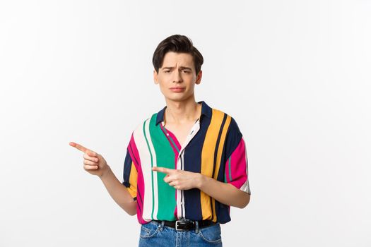 Skeptical gay man looking with disdain and pointing fingers left, disapprove and dislike, standing over white background.